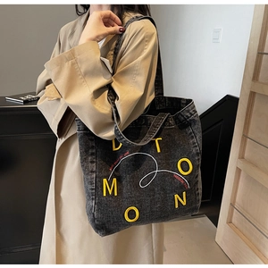 Detail of black denim tote bag with yellow embroidered letters shown on a shoulder