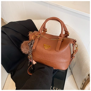 Detail of caramel leather boston bag with metal triangle plaque and plush pom-pom charm