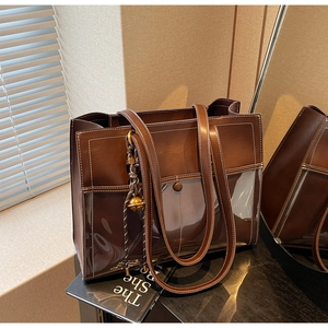 Detail of a brown tote bag featuring transparent PVC panel and a golden bell charm