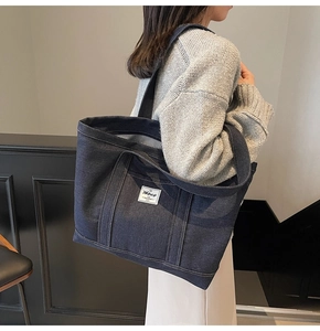 Detail of blue denim tote bag showing the textured fabric, handle, and white logo patch