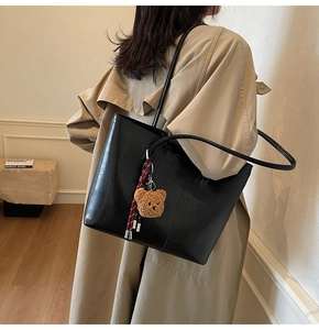 Detailed view of black tote bag featuring a plush bear pendant on a khaki trench coat