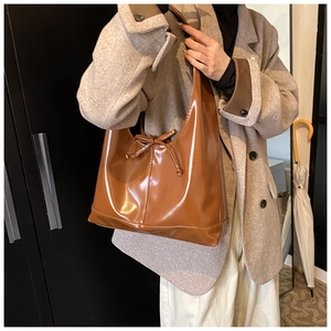 Detail of a large brown glossy shoulder bag with a bow tie accent worn by a model