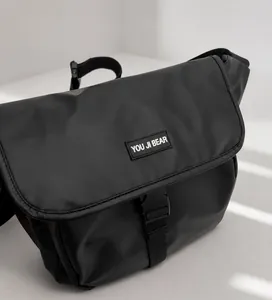 Detail van matzwarte messenger bag met gesp en logo op een denim achtergrond