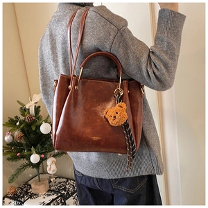 Brown faux leather handbag with plush bear charm worn over the shoulder in cozy home setting