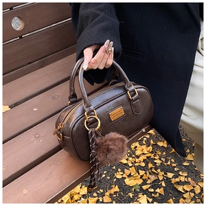 Deep brown Boston bag with gold hardware and pompom on a bench with autumn leaves