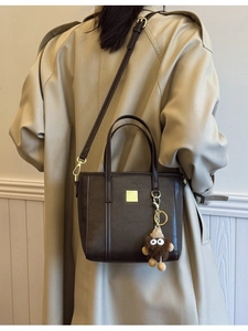 Dark brown faux leather tote bag with gold logo and plush charm worn with trench coat