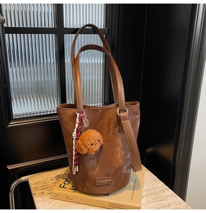 Brown bucket tote bag with teddy dog plush charm and minimalist brand label on surface