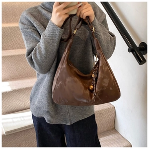 Detail of a brown glossy half-moon hobo bag with a gold bell charm on a model's shoulder