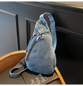 Blue denim crossbody chest bag with adjustable strap and silver zipper on a wooden tray