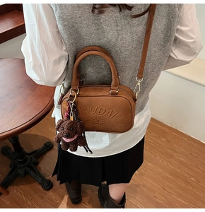 Detailed close-up of a brown suede crossbody bag with embossed letters and a plush toy charm