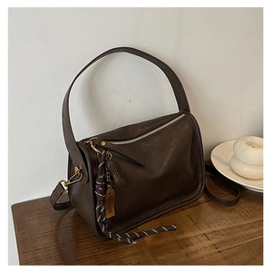 Détail du sac en cuir marron avec pompon tressé et étiquette en métal sur table bois