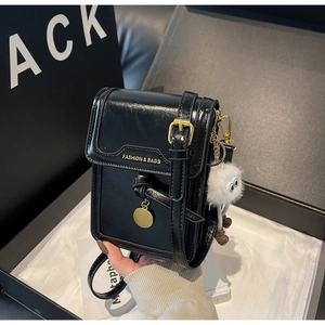 Detail of black crossbody phone bag with glossy texture, gold buckle and plush charm