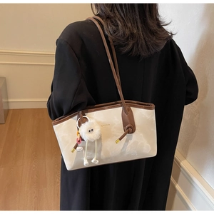 Detail of cream tote bag with brown trim featuring a plush toy charm and gold logo