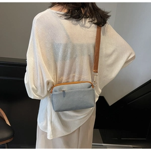 Detail of light blue denim crossbody bag with brown strap and metallic zippers
