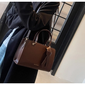 Braune Shopper-Tasche mit dekorativer Schleife und Goldlogo an der Schulter des Models