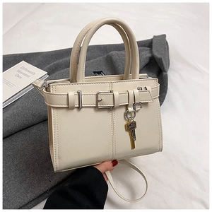 Close-up of cream-colored small square handbag with metal key charm and decorative belt