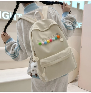 Back view of a cream nylon backpack with colorful pom-poms and a plush monster charm