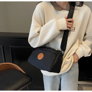 Détail du sac bandoulière noir avec large sangle et écusson rond en cuir marron sur le devant