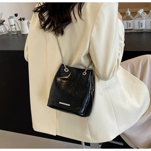 Detail of glossy black bucket bag with silver chain strap worn over a white blazer