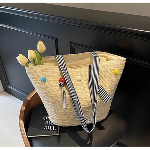 Close-up of woven straw tote bag with colorful plush balls and striped ribbon handle