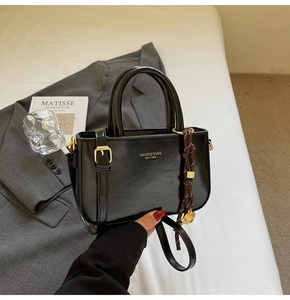 Detail van zwarte glanzende crossbodytas met gouden tekst en bruine geweven hanger