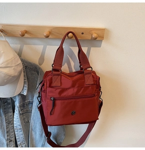 Brick red nylon tote bag detail with zip pocket and metal logo on a wooden hook