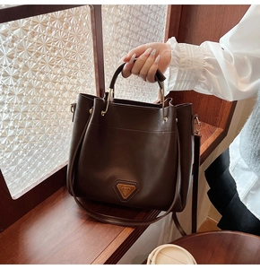 Détail du sac seau marron foncé avec anse dorée et logo métallique sur fond texturé