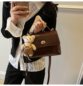 Detalle de bolso bandolera marrón brillante con cierre dorado y colgante de peluche