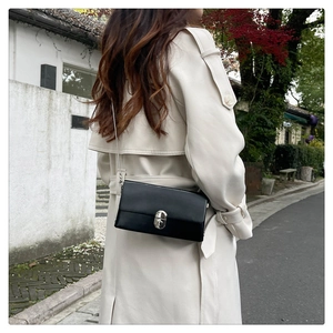 Close-up of a black chain crossbody box bag styled with a beige trench coat outdoors