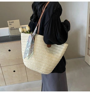 Detail of a woven straw tote bag featuring brown leather handles and a silk scarf accent