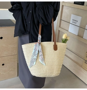 Detail of woven straw tote bag featuring brown leather handles and a floral silk scarf