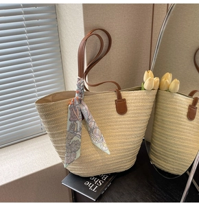 Detail of a beige woven straw tote bag featuring brown leather handles and a blue floral scarf