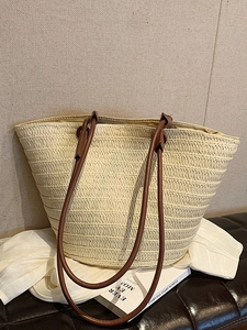 Large beige woven straw tote bag with brown leather handles on a leather bench