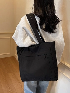 Large black nylon tote bag with front zipper pocket shown on a model in a modern room