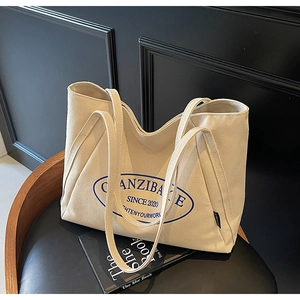 Detalle de bolso tote de lona beige con logo azul y correa resistente sobre silla de cuero