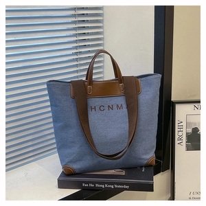 Detail of blue canvas tote bag with brown leather trim and HCNM logo on a stylish desk
