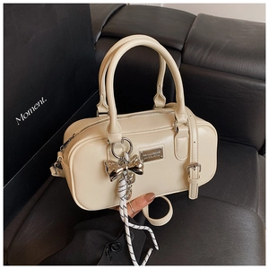 Close-up of beige Boston bag with silver nameplate and bow pendant on leather surface
