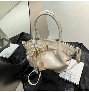 Detail einer cremeweißen Faltenhandtasche mit Perlenspitzen-Bogenanhänger auf Lederjacke