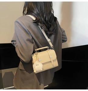 Detail of cream crossbody bag with leather straps, metal buckles, and a fluffy white charm