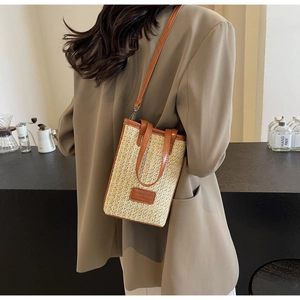Vertical straw woven crossbody bag with brown faux leather trim on a modern café background