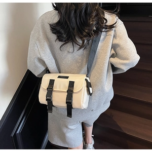 Beige messenger bag with black buckles shown on a model against wooden stairs