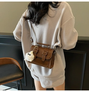 Detail view of a brown leather crossbody bag with flap buckles and white plush pendant accessory