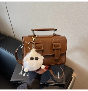 Detail of a brown leather crossbody bag with decorative straps and a plush white pendant