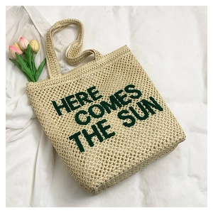 Detail of embroidered 'HERE COMES THE SUN' text on a woven straw tote bag texture