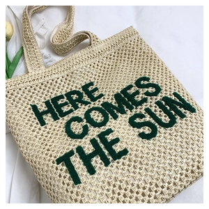 Close-up of embroidered Here Comes The Sun text on beige woven straw beach tote bag