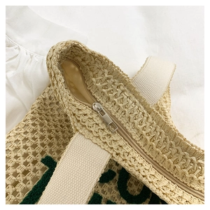 Detail of woven straw tote bag with green tufted letters, metal zipper and canvas strap
