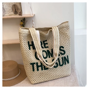 Detail of a beige woven tote bag with green Here Comes The Sun embroidery and straw hat