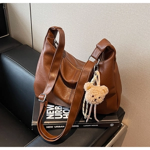 Brown faux leather tote bag with dual straps and plush bear charm on a black sofa