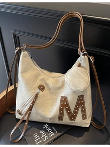 Beige tote bag with brown M logo and silver studs on a dark chair with a small pouch