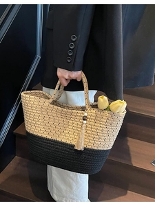 Woven straw tote bag featuring black and beige color-block design with tassel detail
